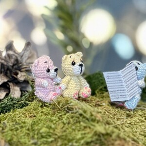 May include: Three small, crocheted teddy bears in pink, yellow, and blue, sitting on a bed of green moss. One bear holds a small sheet of paper. A pine cone is visible in the background.