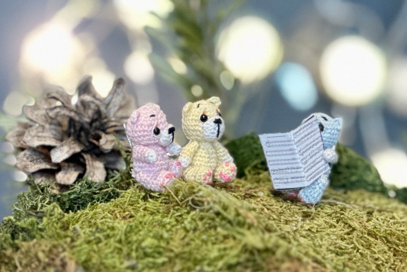 May include: Three small, crocheted teddy bears in pink, yellow, and blue, sitting on a bed of green moss. One bear holds a small sheet of paper. A pine cone is visible in the background.