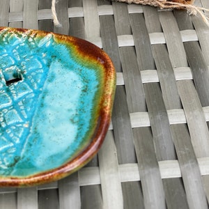 May include: A turquoise blue ceramic soap dish with a brown rim. The dish has a textured surface and a small hole in the center.