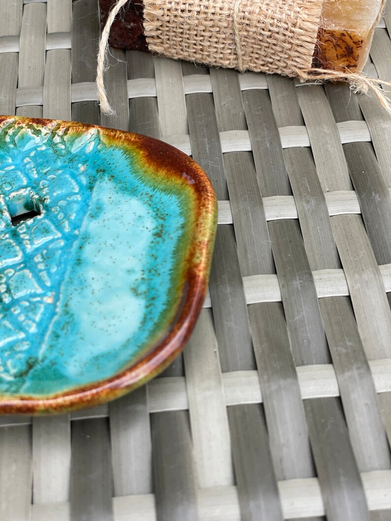 May include: A turquoise blue ceramic soap dish with a brown rim. The dish has a textured surface and a small hole in the center.