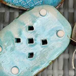 May include: A light blue ceramic soap dish with a decorative design of small squares. The dish has four white feet on the bottom and is designed to drain water.