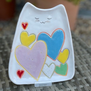 May include: White ceramic cat-shaped dish with a hand-painted design of colorful hearts. The cat has closed eyes and a small mustache.