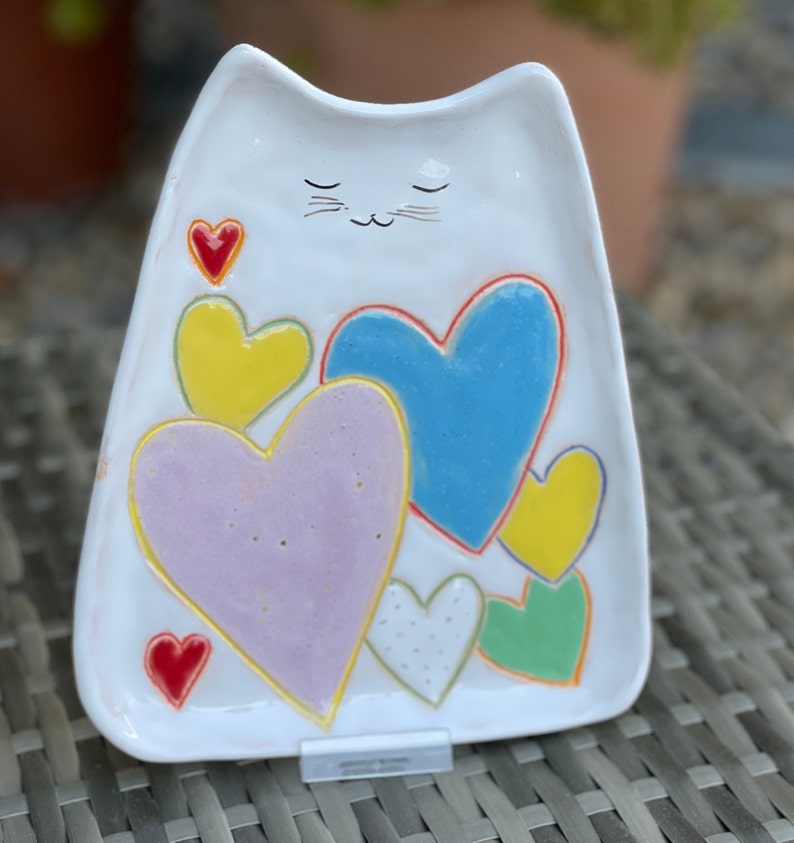 May include: White ceramic cat-shaped dish with a hand-painted design of colorful hearts. The cat has closed eyes and a small mustache.