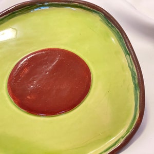 May include: A ceramic avocado-shaped serving dish with a green exterior and a brown interior. The dish has a raised rim and a smooth, glossy finish.