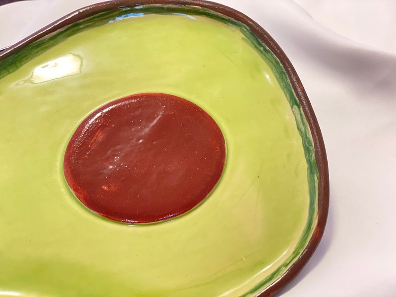 May include: A ceramic avocado-shaped serving dish with a green exterior and a brown interior. The dish has a raised rim and a smooth, glossy finish.