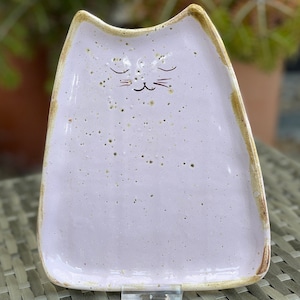 May include: A light purple ceramic cat-shaped dish with a speckled glaze and a hand-painted face. The dish has a slightly raised rim and a flat bottom.