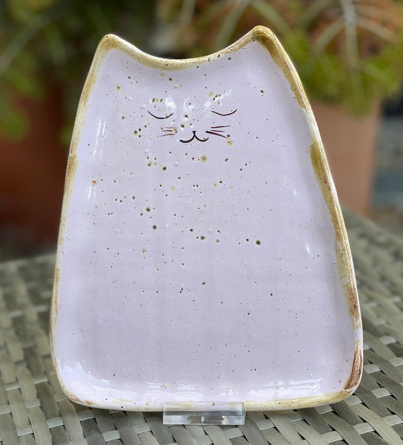 May include: A light purple ceramic cat-shaped dish with a speckled glaze and a hand-painted face. The dish has a slightly raised rim and a flat bottom.