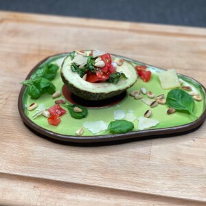 May include: A halved avocado filled with a salad of chopped tomatoes, basil, and pine nuts, topped with grated parmesan cheese. The avocado is served on a green ceramic plate with additional pine nuts, basil, and parmesan cheese scattered around it.