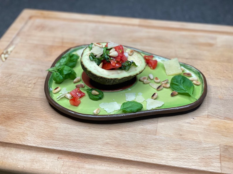 May include: A halved avocado filled with a salad of chopped tomatoes, basil, and pine nuts, topped with grated parmesan cheese. The avocado is served on a green ceramic plate with additional pine nuts, basil, and parmesan cheese scattered around it.