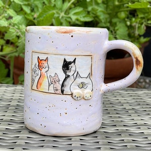 Peut inclure: Une tasse en céramique lavande avec un motif peint à la main de quatre chats. Les chats sont dessinés dans un style cartoon et regardent par une fenêtre. La tasse a un glaçage moucheté et une poignée arrondie.