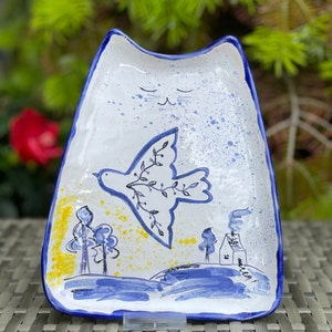 May include: A white ceramic dish shaped like a cat with a blue rim. The dish has a hand-painted design of a dove with an olive branch flying over a blue and yellow landscape with trees and a house.