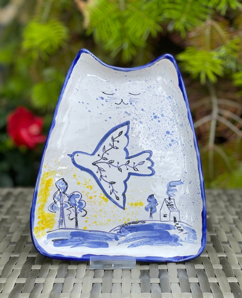 May include: A white ceramic dish shaped like a cat with a blue rim. The dish has a hand-painted design of a dove with an olive branch flying over a blue and yellow landscape with trees and a house.