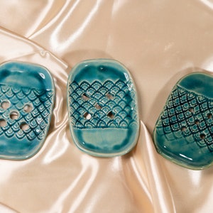 May include: Three teal blue ceramic soap dishes with a decorative pattern. Each dish has a different pattern, but all have a raised design.