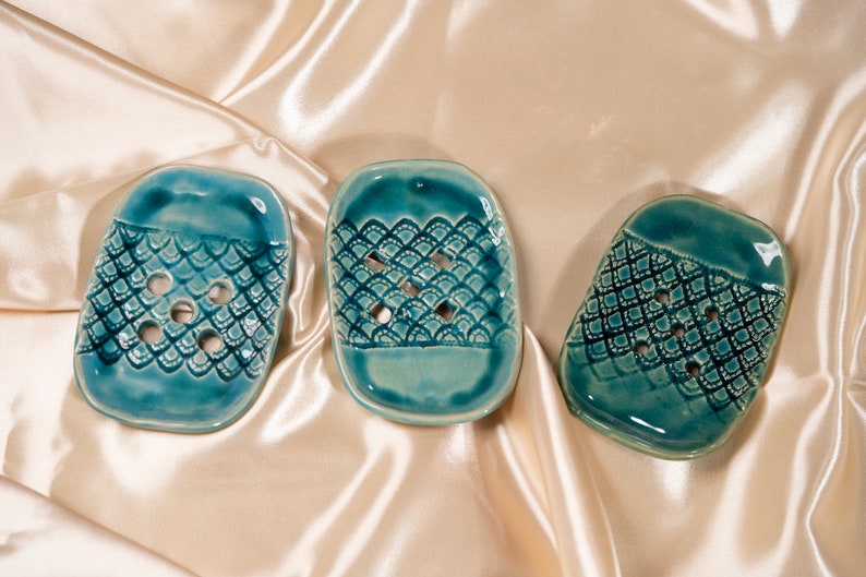 May include: Three teal blue ceramic soap dishes with a decorative pattern. Each dish has a different pattern, but all have a raised design.