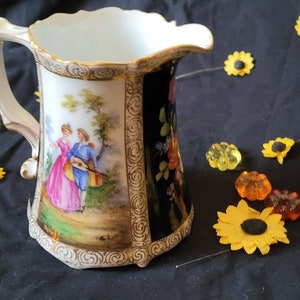May include: A white porcelain pitcher with a black and gold floral design. The pitcher has a handle and a spout. The pitcher is decorated with a scene of a couple in a garden.