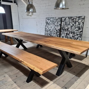 Könnte beinhalten: Ein langer Holztisch mit lebenden Kanten und schwarzen X-förmigen Beinen. Eine passende Holzbank mit schwarzen X-förmigen Beinen steht vor dem Tisch.