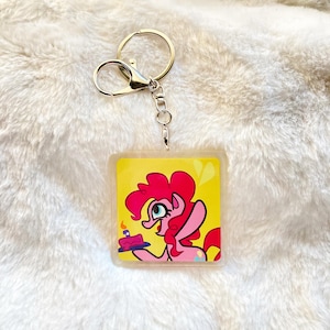 Puede incluir: Un llavero cuadrado con una imagen de dibujos animados de un pony rosa sosteniendo un pastel de cumpleaños sobre un fondo amarillo. El llavero tiene un cierre y un anillo de metal plateado. El pony tiene una melena y una cola rosas.