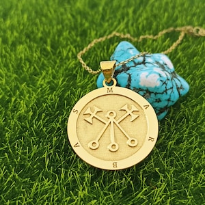 May include: A gold necklace with a round pendant featuring a symbol with three crossed spears and the letters "M A S A B R" around the edge.