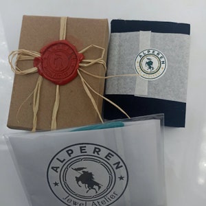 May include: A brown box with a red wax seal and a beige ribbon. A black box with a white label that says "Alperen Jewel Atelier" and a green and white logo. A white bag with a black label that says "Alperen Jewel Atelier" and a green and white logo.