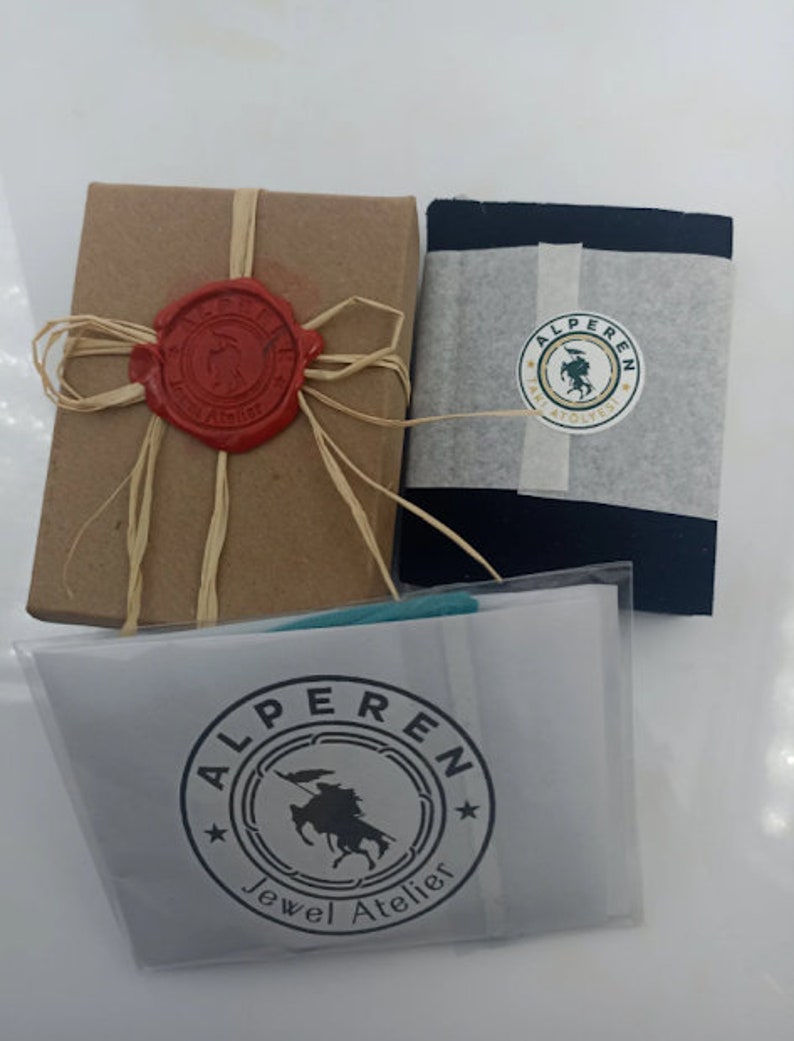 May include: A brown box with a red wax seal and a beige ribbon. A black box with a white label that says "Alperen Jewel Atelier" and a green and white logo. A white bag with a black label that says "Alperen Jewel Atelier" and a green and white logo.