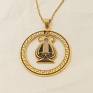May include: Gold necklace with a circular pendant featuring a black lyre design. The pendant has a Greek key border.