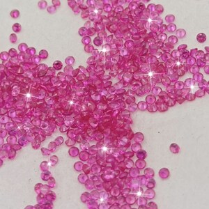 Peut inclure: Un grand groupe de pierres pr&eacute;cieuses roses, &eacute;parpill&eacute;es sur une surface blanche. Les pierres pr&eacute;cieuses sont rondes et ont une finition brillante et facet&eacute;e.