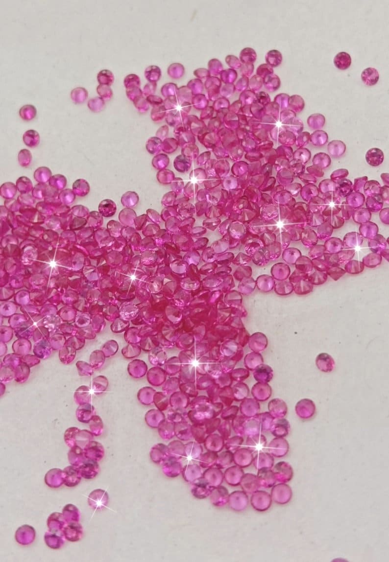 Peut inclure: Un grand groupe de pierres pr&eacute;cieuses roses, &eacute;parpill&eacute;es sur une surface blanche. Les pierres pr&eacute;cieuses sont rondes et ont une finition brillante et facet&eacute;e.