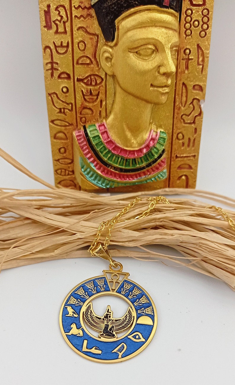 May include: Gold necklace with a blue and gold pendant featuring an Egyptian goddess with outstretched wings. The pendant is surrounded by hieroglyphs and a sun disk.