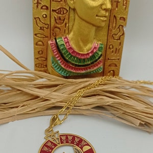 May include: Gold necklace with a red and gold pendant featuring an Egyptian goddess with wings. The pendant is surrounded by hieroglyphs and a sun disk.