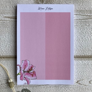 Puede incluir: Un bloc de notas rosa y blanco con el texto "Meine Notizen" en la parte superior. El bloc de notas tiene una ilustración de flor de lirio rosa en la esquina inferior izquierda.