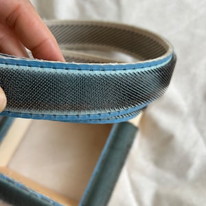 May include: A close-up of a blue and white fabric brush with metal bristles. The brush is rolled up and held in a hand. The brush is designed for cleaning and grooming.