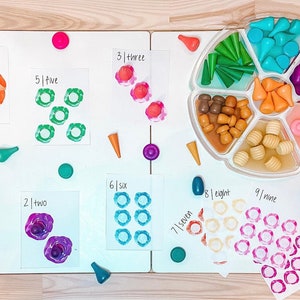 May include: A set of ten colorful cards with numbers 1 through 10 written on them. Each card has a different color and a different number of circles. The circles are painted in different colors. There are also small wooden pieces in different colors and shapes on the table.