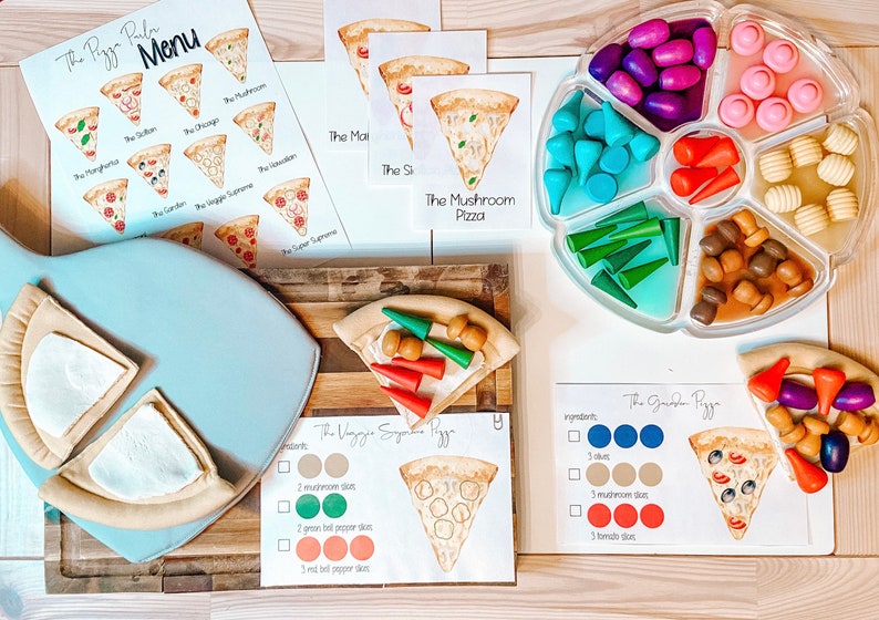 May include: A pizza making play set with a menu, pizza slices, and toppings. The menu includes different pizza options like "The Veggie Supreme" and "The Garden Pizza". The toppings are made of wood and include mushrooms, peppers, and olives.