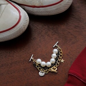 May include: A multi-strand bracelet with faux pearls, gold and black chains, and a smiley face charm. The bracelet is next to a pair of white canvas sneakers with red trim and a red garment on a wooden surface.