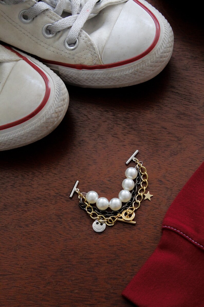 May include: A multi-strand bracelet with faux pearls, gold and black chains, and a smiley face charm. The bracelet is next to a pair of white canvas sneakers with red trim and a red garment on a wooden surface.