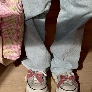 May include: A pair of white platform sneakers with brown soles and pink velvet bows. The sneakers are worn with light blue jeans. A pink and beige woven bag is visible on the left side of the image.
