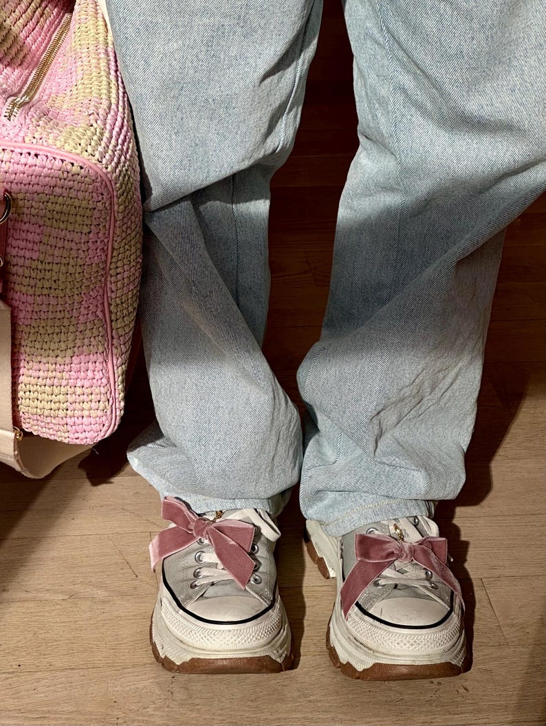 May include: A pair of white platform sneakers with brown soles and pink velvet bows. The sneakers are worn with light blue jeans. A pink and beige woven bag is visible on the left side of the image.