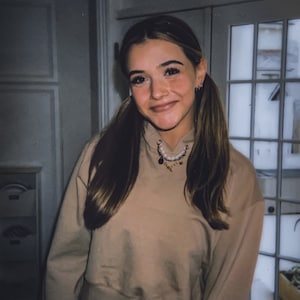May include: A young person with long brown hair in pigtails smiles, wearing a tan cropped hoodie and blue jeans. A necklace with charms hangs around the neck. The background includes a white door with glass panes and a white wall.
