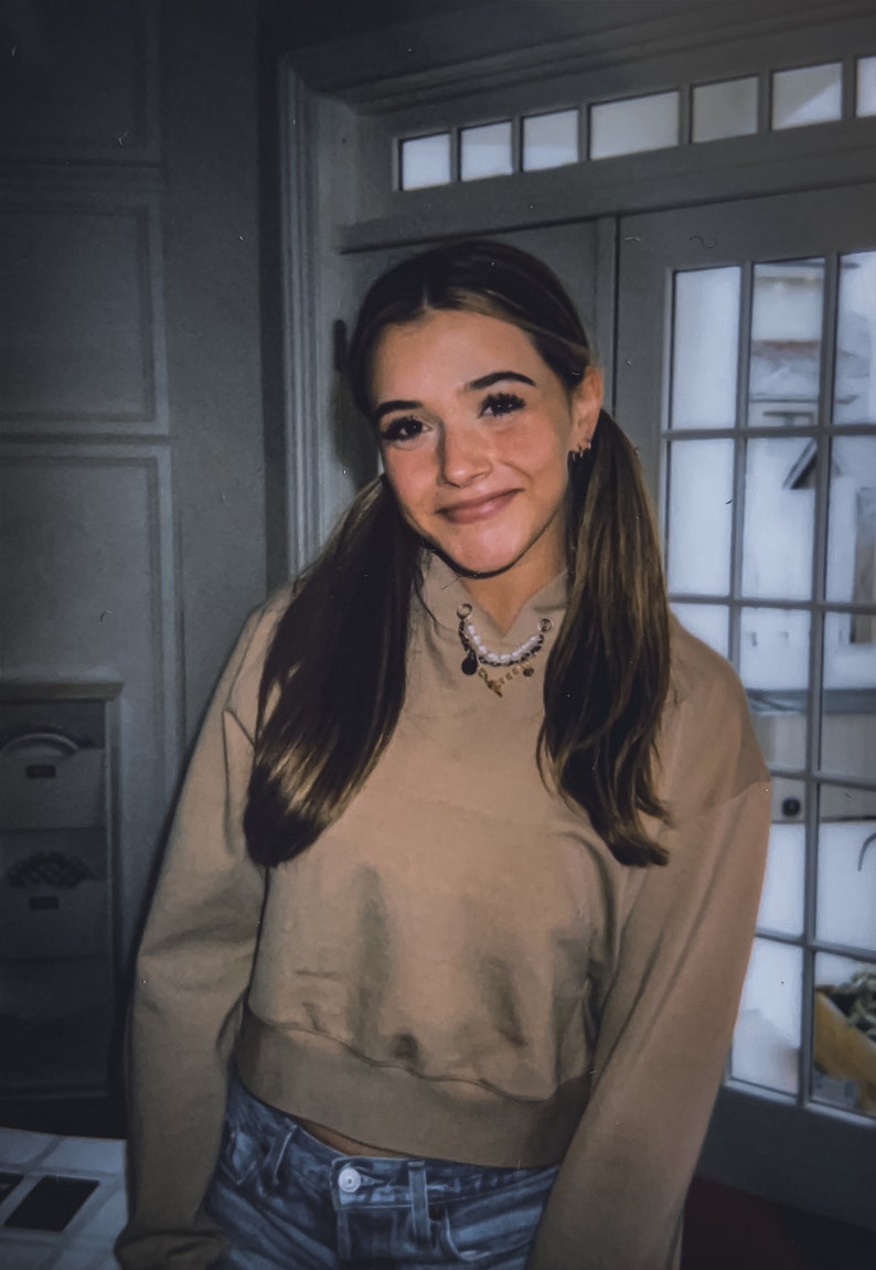 May include: A young person with long brown hair in pigtails smiles, wearing a tan cropped hoodie and blue jeans. A necklace with charms hangs around the neck. The background includes a white door with glass panes and a white wall.
