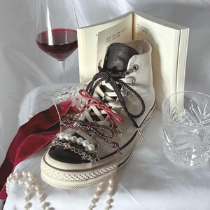 May include: Cream-colored high-top sneaker with black trim, adorned with pearl and chain embellishments, and red and brown laces. A glass of red wine, a book, and a decorative glass are in the background. A pearl necklace is draped in front.