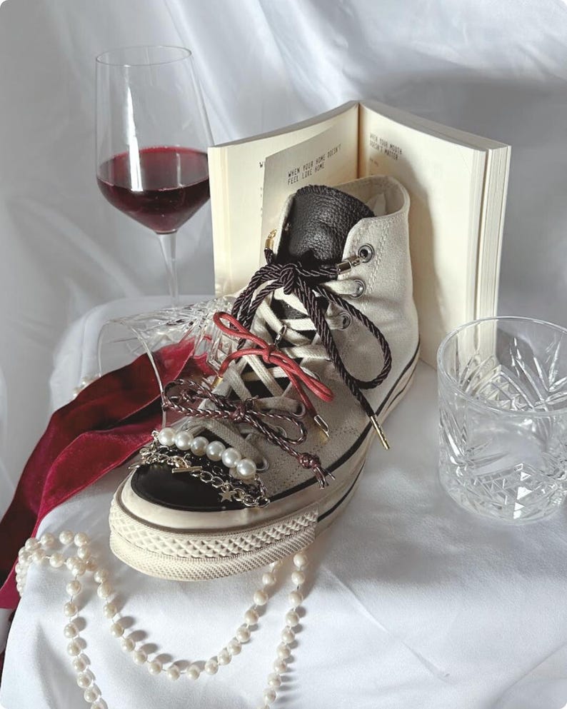 May include: Cream-colored high-top sneaker with black trim, adorned with pearl and chain embellishments, and red and brown laces. A glass of red wine, a book, and a decorative glass are in the background. A pearl necklace is draped in front.