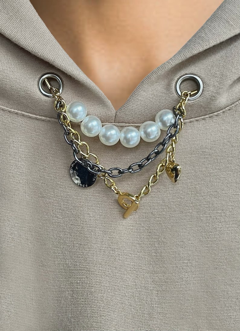 May include: A layered necklace with faux pearls and gold and silver chains. The necklace has a coin charm, a heart charm, and a charm with the letter "P". The necklace is attached to a tan hoodie.