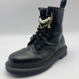 May include: Black leather combat boot with a pearl and gold chain accessory. The boot features a lace-up design and a thick, black sole. A small skull charm hangs from the chain.