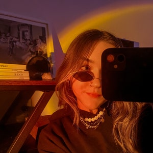 May include: A person wearing oval sunglasses and a gold chain necklace with pearls and charms. The person is holding a black smartphone, partially obscuring their face. A shelf with books and a dark glass bottle is visible in the background.