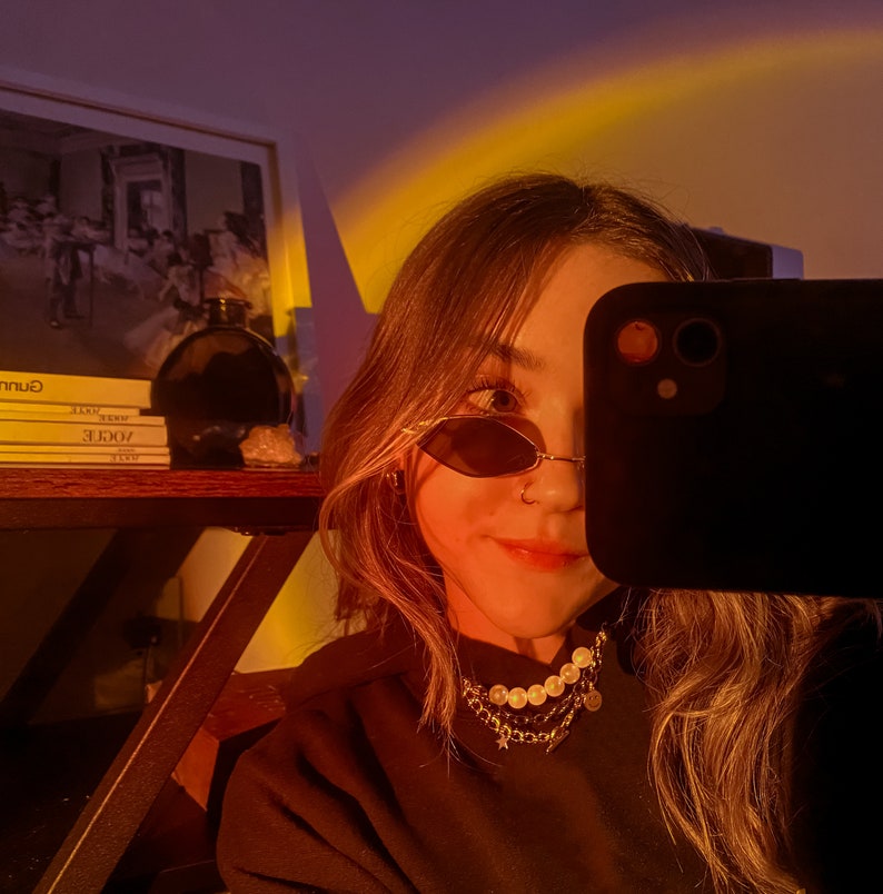 May include: A person wearing oval sunglasses and a gold chain necklace with pearls and charms. The person is holding a black smartphone, partially obscuring their face. A shelf with books and a dark glass bottle is visible in the background.