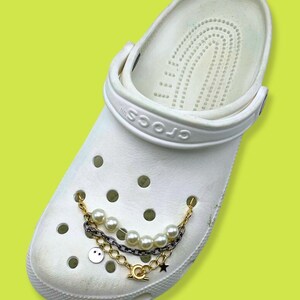 May include: White Crocs shoe with a decorative chain featuring faux pearls, a smiley face charm, and a star charm. The chain is attached to the shoe with gold-colored and silver-colored chains.
