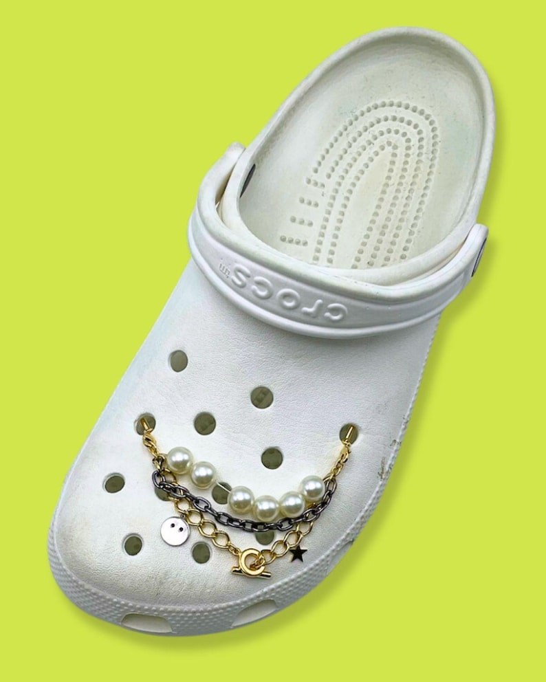 May include: White Crocs shoe with a decorative chain featuring faux pearls, a smiley face charm, and a star charm. The chain is attached to the shoe with gold-colored and silver-colored chains.