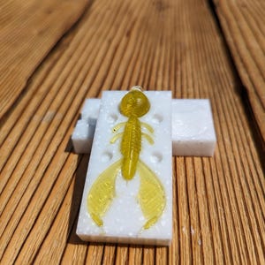 May include: A white plastic mold with a yellow plastic lure inside. The lure is shaped like a shrimp with a segmented body and two large, fan-shaped appendages.