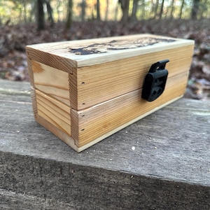 May include: A rectangular wooden box with a black clasp. The box is made of light-colored wood with visible grain and a darker design on the top. The box is sitting on a weathered wooden surface.