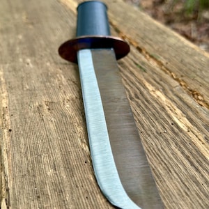 May include: A fixed-blade knife featuring a dark, cylindrical handle and a polished metal blade with a distinct, darkened finish. A circular, copper-toned guard separates the handle from the blade, which tapers to a sharp, curved point.